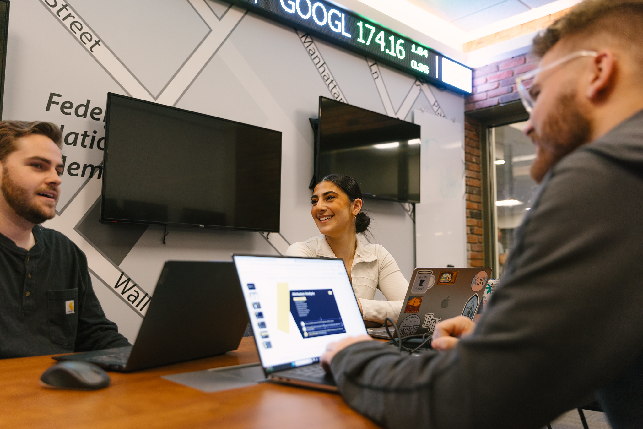 Three students in a room with screens on the wall discussing a project, looking at laptops, and smiling.