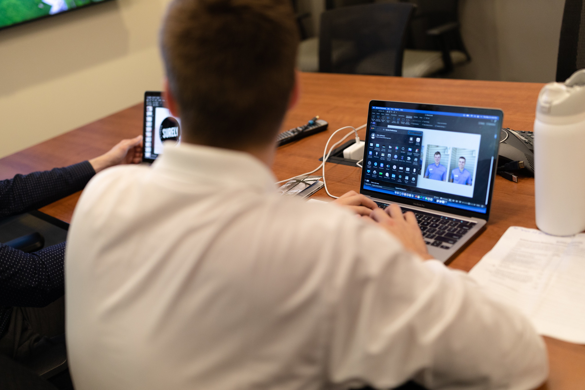 Students collaborating in a conference room, working on laptops and a tablet during a presentation.